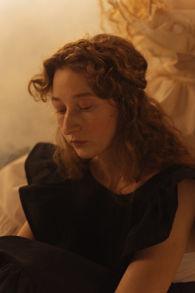 Young woman with curly hair wearing a black top or dress looking down in sadness. 