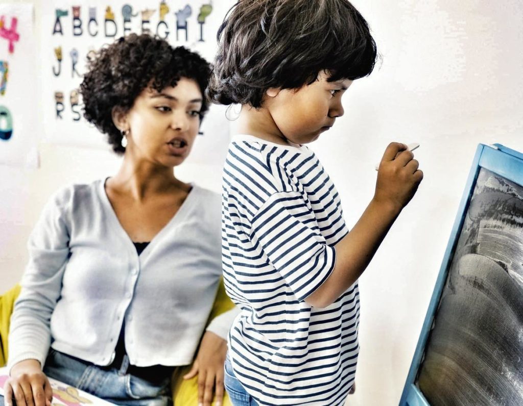 Teacher sitting in a yellow chair with the ABCs behind her on the wall. She is talking to a little boy in a striped shirt who is standing at a blackboard with a look of deep concentration on his face. 