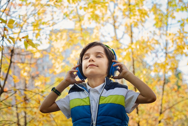Child outside with blue headphones on his ears. He wears a colorful green, blue, black, and white puffy vest and stands in front of fall trees. 