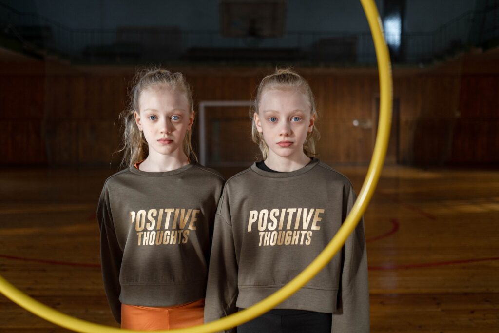 Twin girls in gym class with blonde hair wearing matching sweatshirts that read, "Positive Thoughts". They're staring intently into the camera through a yellow hoop. 