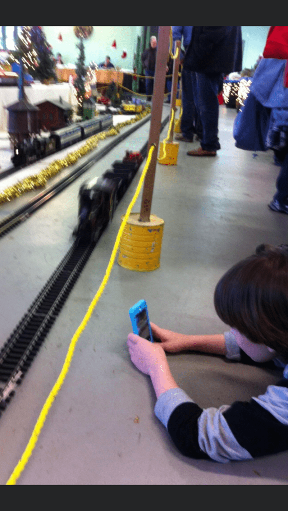 A little boy laying on his stomach taking a photo of a train track with his phone. 