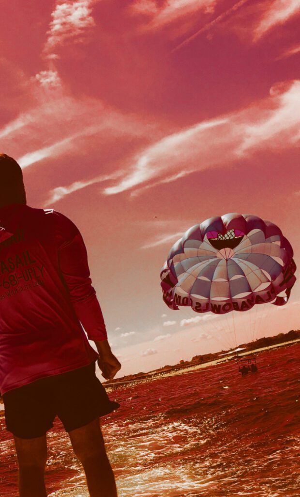 Stylized photo of a little boy standing by the ocean with a colorful parasail in the background. 