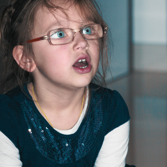 Photo of a child with glasses and bright blue eyes staring up with her mouth open as if she's talking to someone off-camera