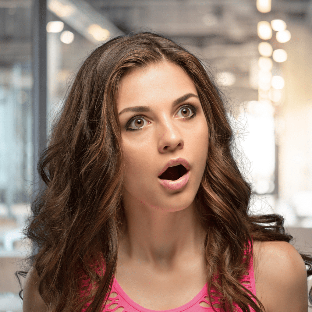 Woman with a pink top and long, light brown hair stands in an office, staring at someone off-camera with wide eyes and an open mouth