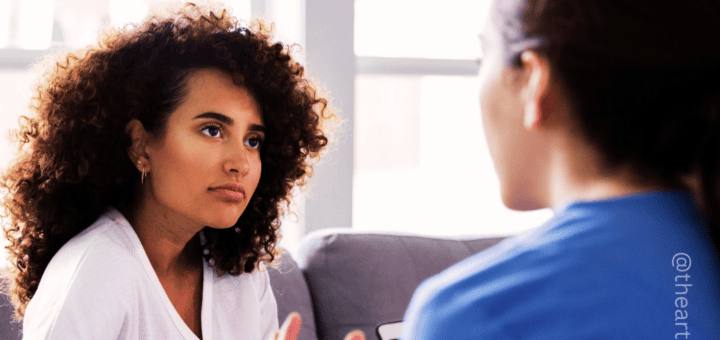 Photo of two women talking. One is front-facing with curly hair and a white top. The other is looking at the first woman. She has dark hair and is wearing a blue shirt