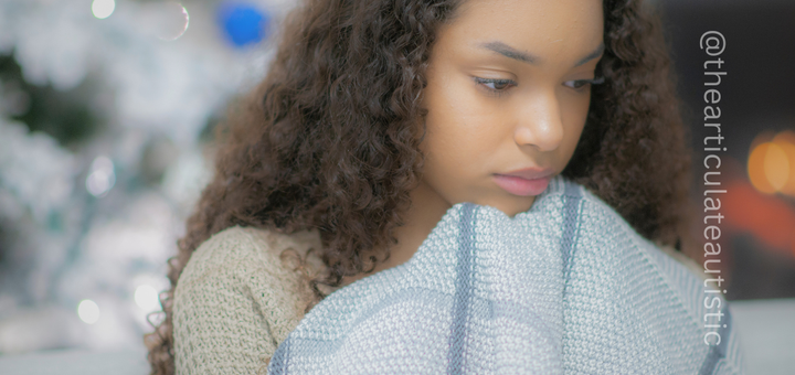 Young girl with long brown, curly hair sits on a couch holding a light blue blanket up to her chin. She looks down and has a faraway or sad look in her eyes. Behind her is a Christmas tree and a fireplace